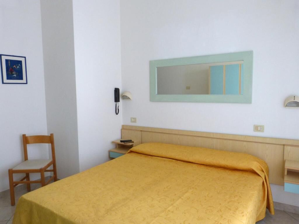 A room with a bed, a chair, a bedside table, a mirror, and a wall-mounted phone.