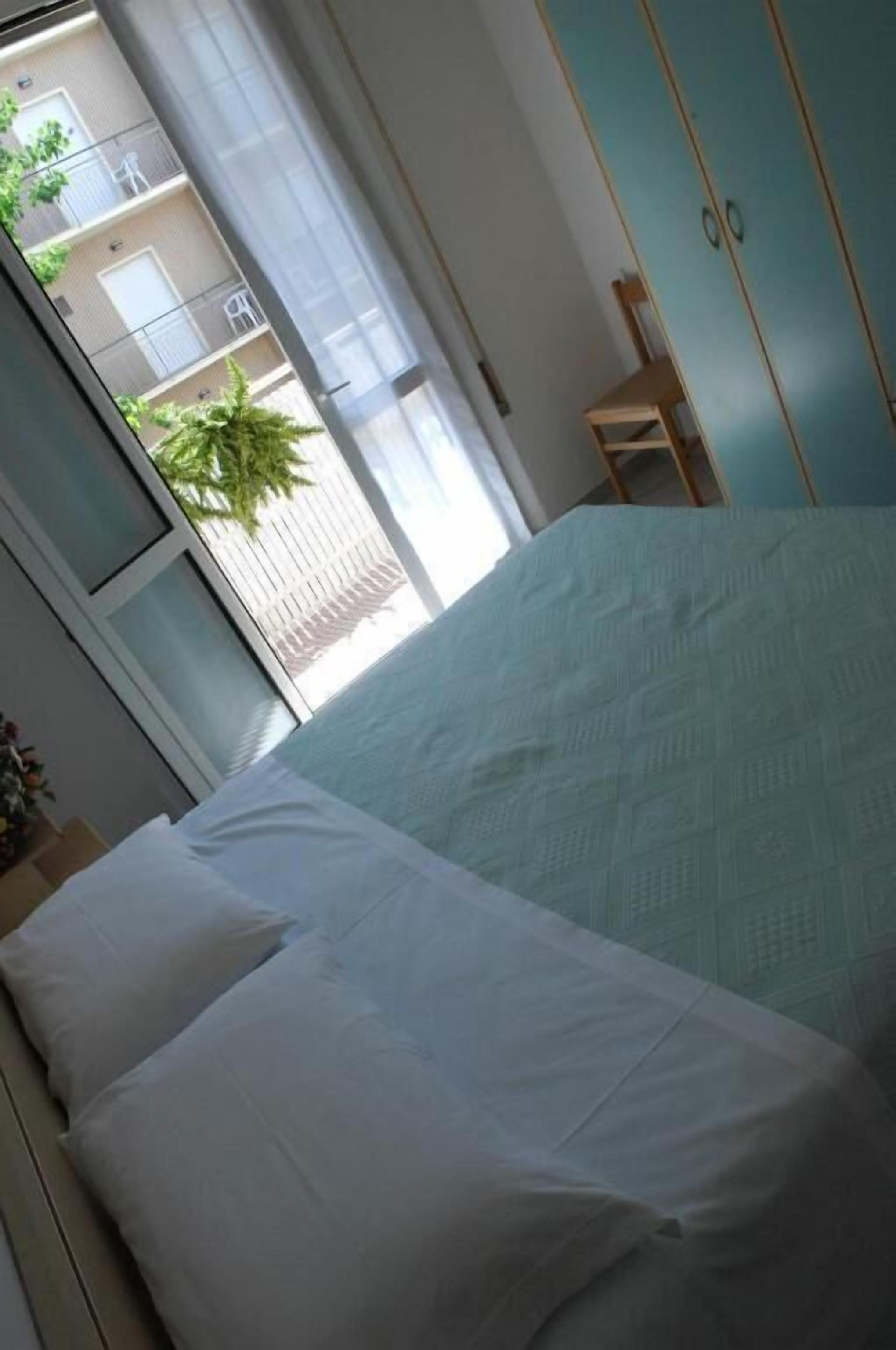 Una cama con ropa de cama blanca, una ventana con cortinas traslúcidas y vistas a un balcón con plantas, además de un armario color turquesa.
