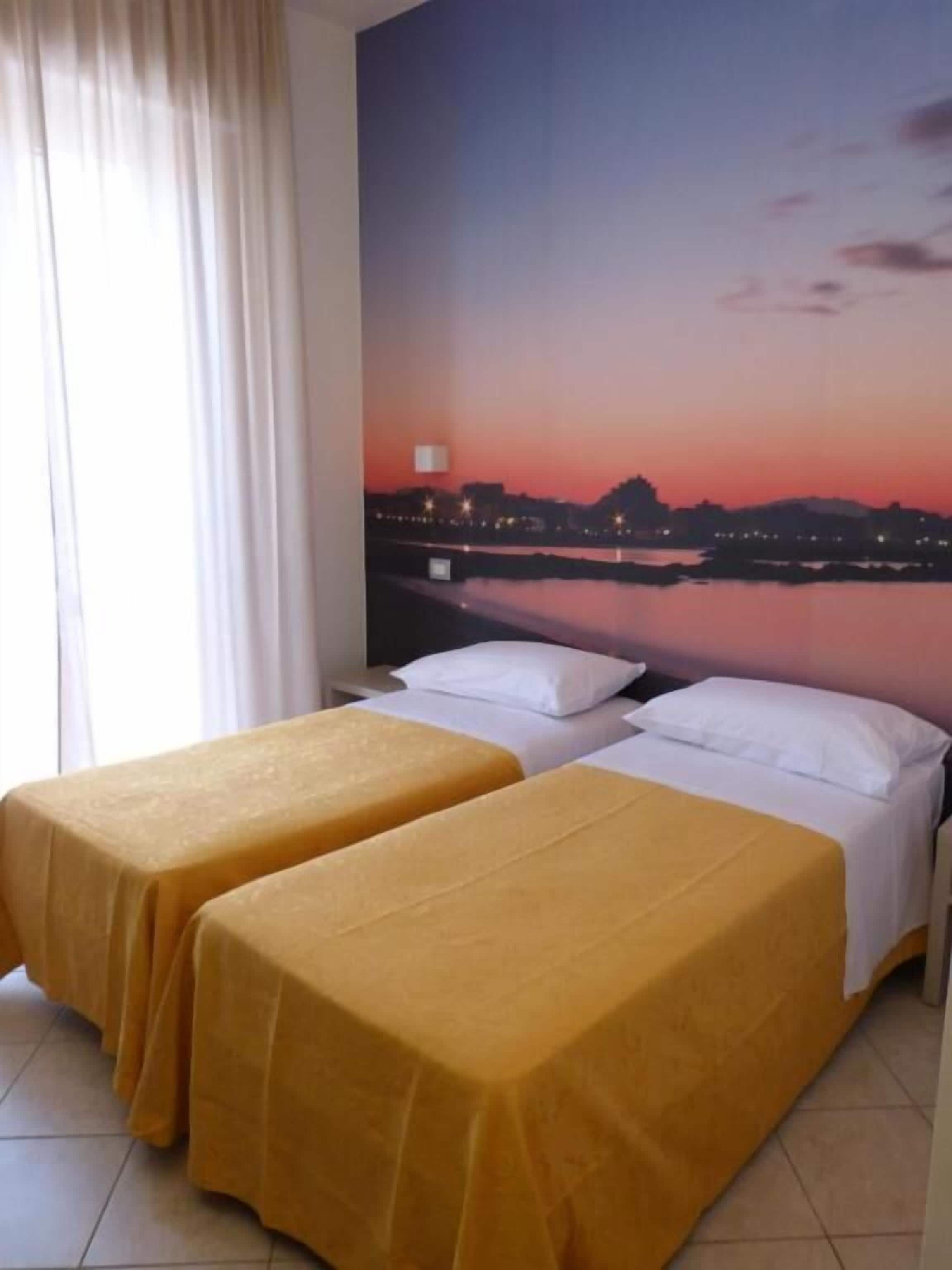 Habitación de hotel con dos camas individuales, un ventanal con cortinas y un mural de paisaje urbano en la pared.