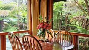 A wooden dining table with six chairs, a vase with flowers, and a view of a lush green forest through large windows. - The Pacifica Inn/ The Volcano Room (Volcano)
