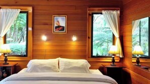 A wooden cabin room with a bed, two windows with curtains, a wall-mounted picture, and two bedside lamps. - The Pacifica Inn/ The Volcano Room (Volcano)
