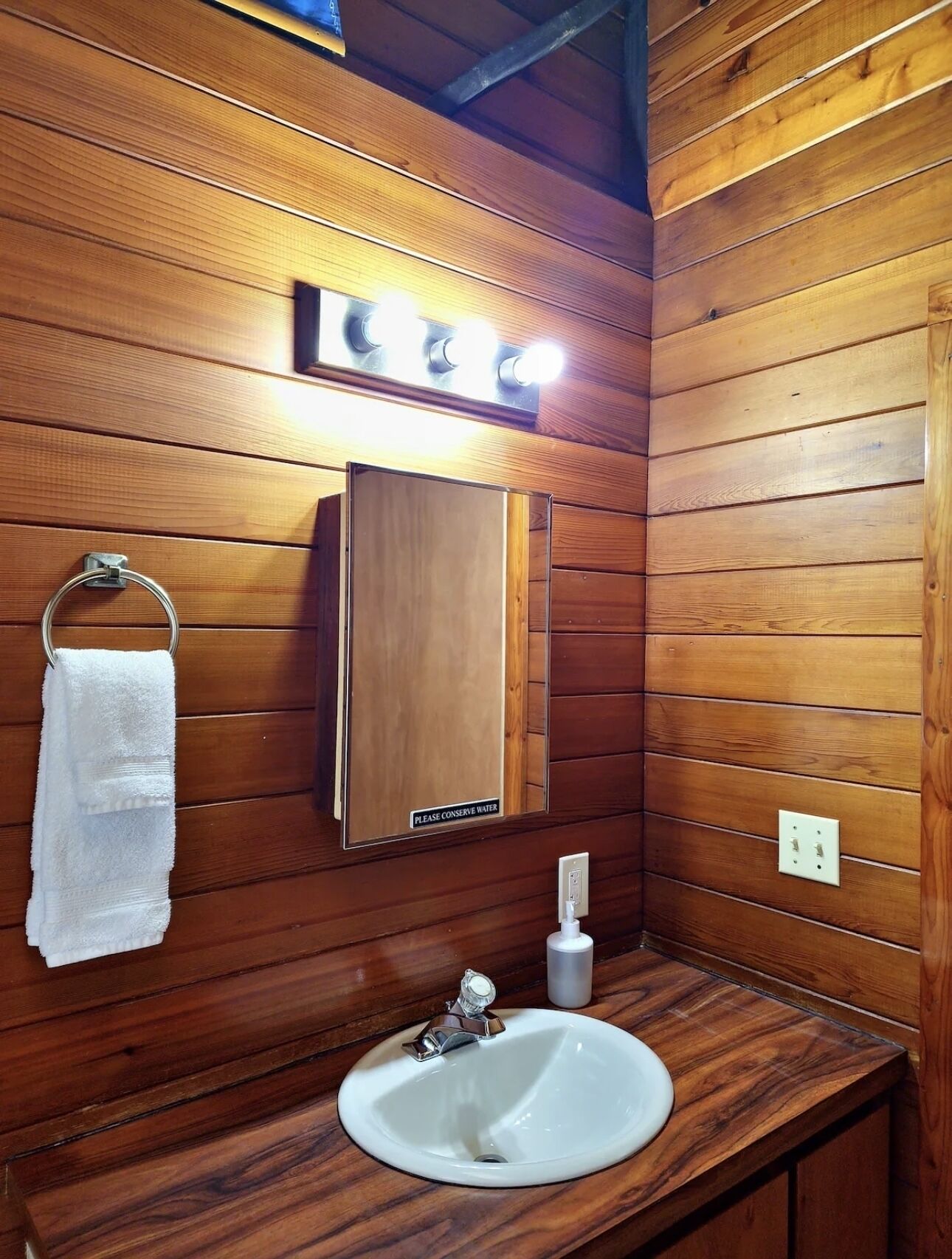 Un baño con paneles de madera, un lavamanos, un perchero para toallas, un espejo y una lámpara.