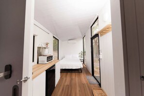 Un couloir étroit mène à une chambre avec un lit, une petite kitchenette équipée d’un four à micro-ondes et une fenêtre.