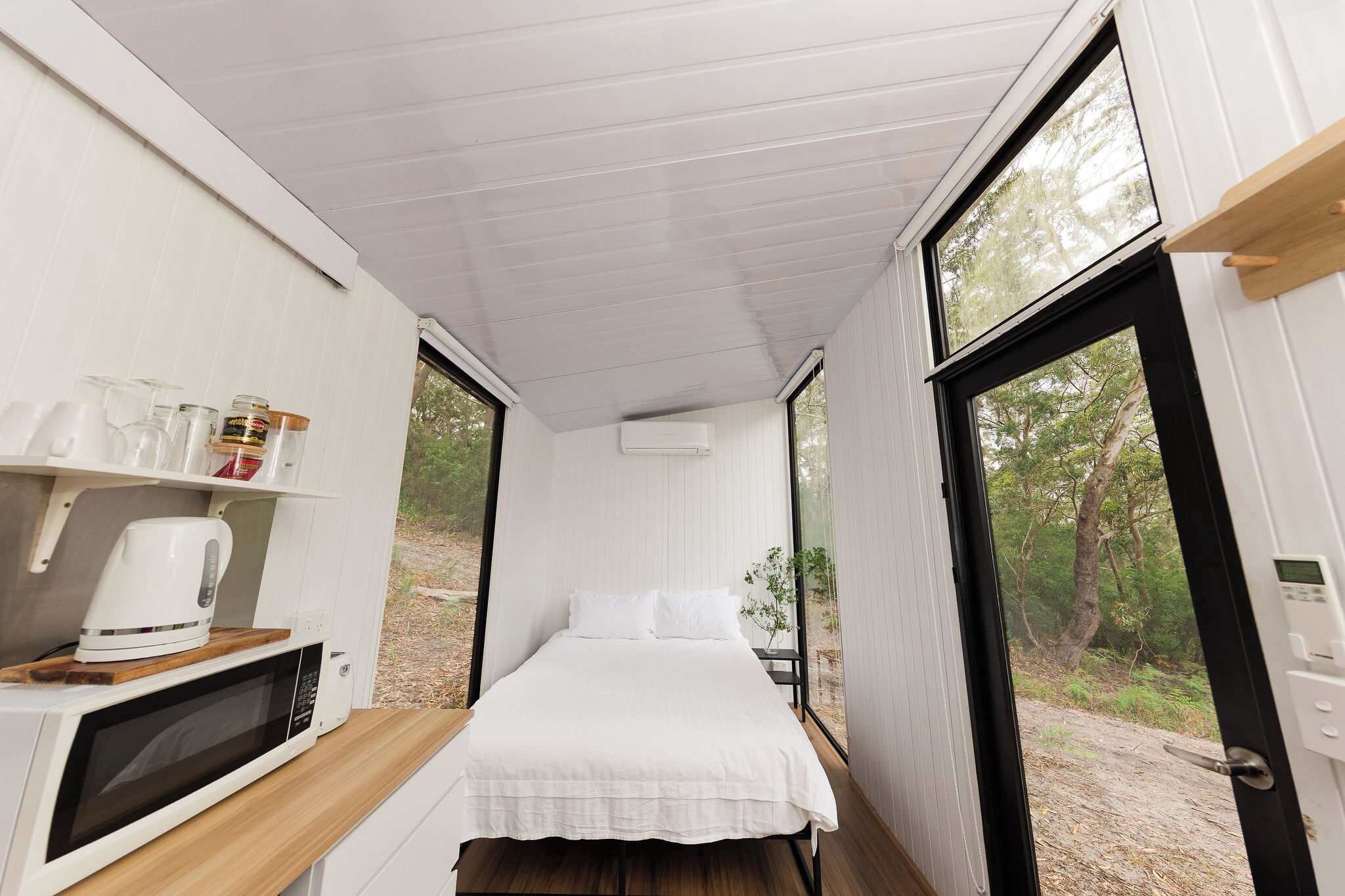 A compact bedroom with a bed, a small table with a kettle, and a window providing a view of the outdoors.