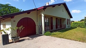 Una casa de dues plantes amb una porta vermella, un balcó i un jardí ben cuidat.