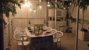 Zona de menjador a l'aire lliure amb taula preparada per a sis persones, envoltada de cadires blanques. Hi ha ampolles i vasos sobre la taula, i una petita taula de fusta. La zona està decorada amb llums de cadena i vegetació.