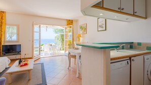 Une pièce comprenant une télévision, une table à manger, un canapé, un petit coin cuisine avec un évier et un lave-linge, et offrant une vue sur la mer par une porte ouverte.