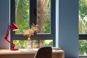 A desk with a red desk lamp, a vase with dried flowers, and a view of greenery through the window.