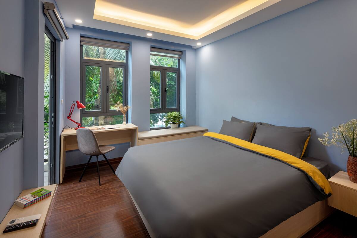 A bedroom with a large bed, a desk with a chair, and a window with a view of greenery.