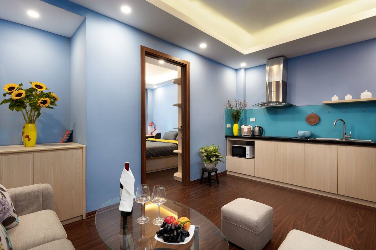 A modern hotel room with a blue wall, wooden floor, a glass table with a fruit platter, wine glasses, and a vase of sunflowers.