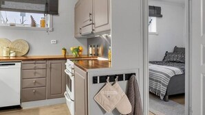 A compact kitchenette with a stove, oven, and a small countertop. There is a window with a blind, and a bed with a striped blanket in the adjacent room.