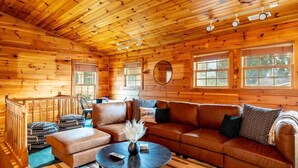 A cozy cabin interior with a wooden ceiling, a large brown leather sofa, a round coffee table, and a dining area with a table and chairs. - Serene Cabin, Sleeps 12 W Views・Hot tub・Games (Sevierville)