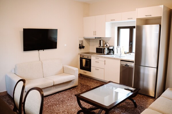 A compact living space with a sofa, a glass-top coffee table, a flat-screen TV mounted on the wall, a kitchenette with a microwave, oven, and refrigerator, and a window providing natural light.