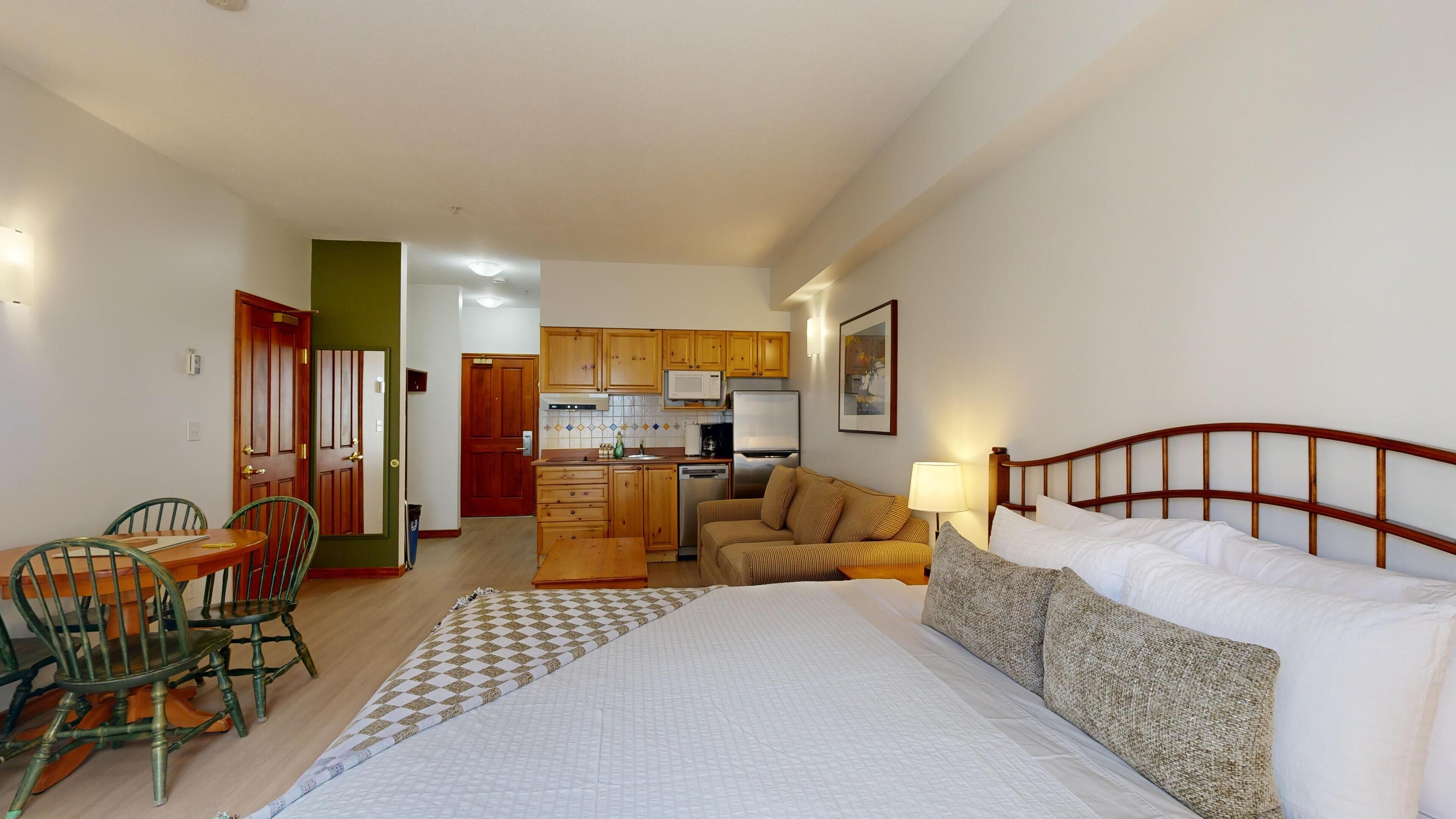 A compact hotel room with a bed, a sofa, a dining table with chairs, a kitchenette, and a bathroom.