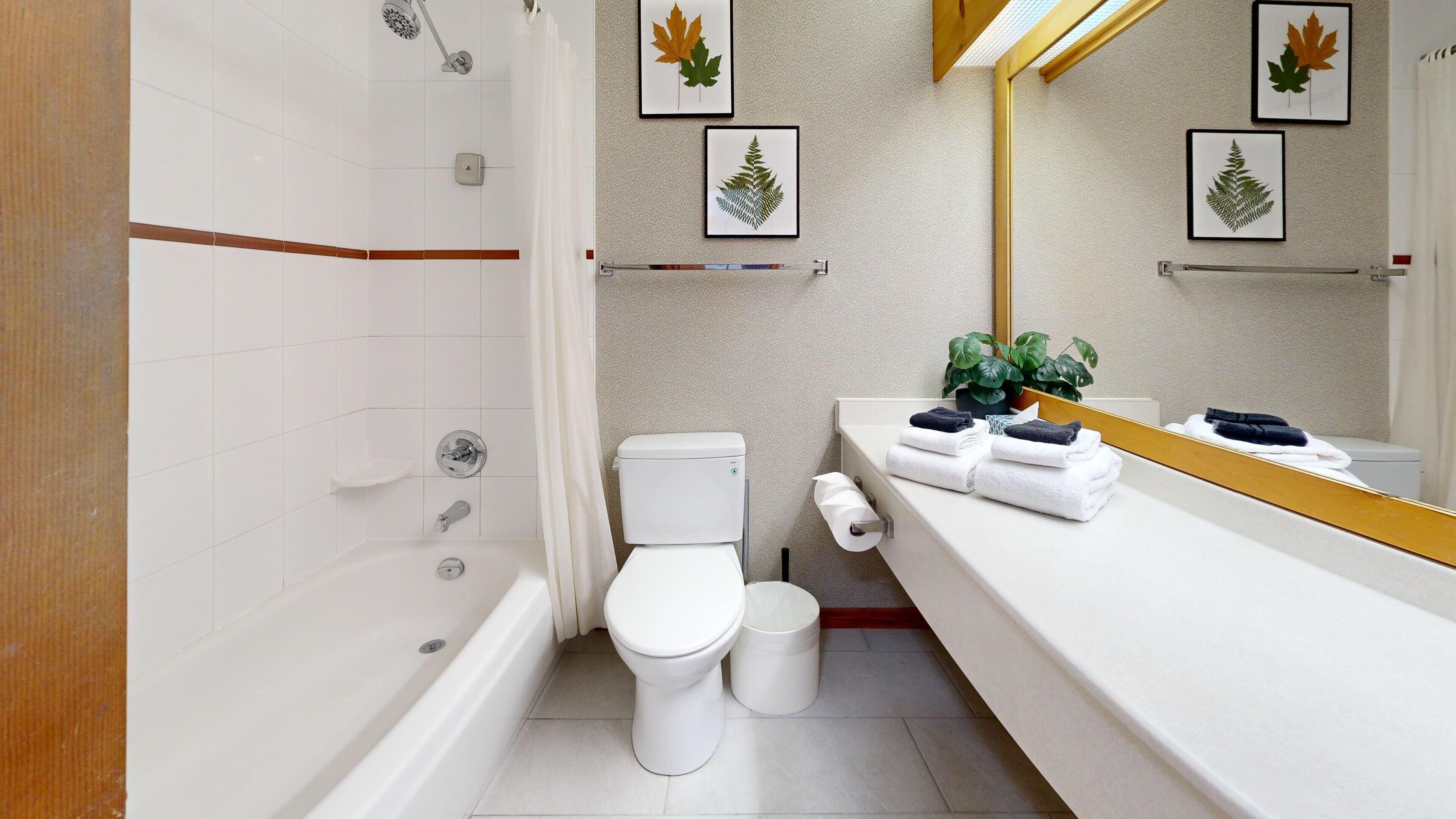 Een moderne badkamer met bad, toilet en wastafel. Er hangen kunstwerken met bladeren in lijsten aan de muur, een potplant en handdoeken op de wastafel.