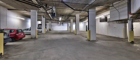 A parking garage with concrete floors and pillars, fluorescent lighting, and visible pipes and ducts.