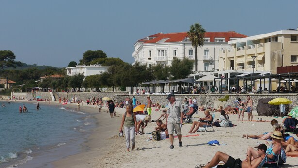 Une plage où des gens se prélassent au soleil et se promènent le long du rivage. On aperçoit des bâtiments à l’arrière-plan et le ciel est dégagé.