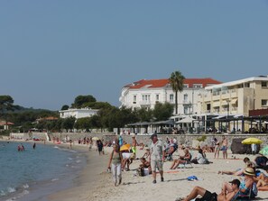 A beach with people sunbathing and walking along the shore. There are buildings in the background and a clear sky.