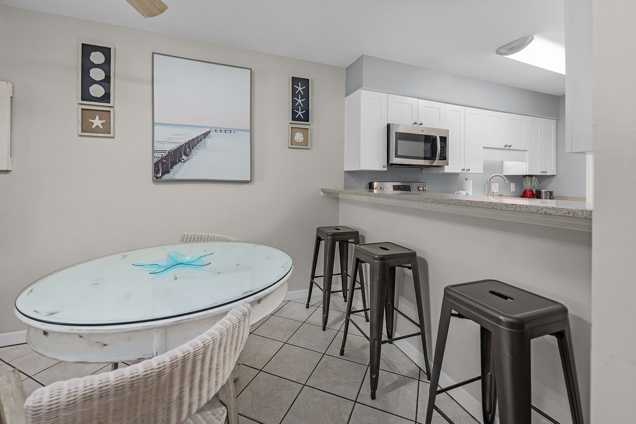 Een keuken met witte kasten, een magnetron en een spoelbak. Er staat een glazen tafel met een zeester decoratie en twee krukken.