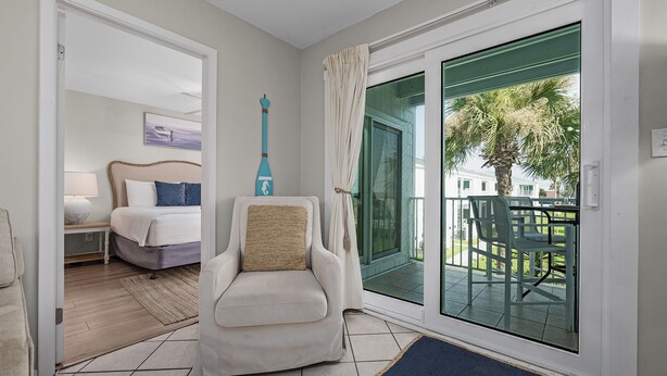 A hotel room with a bed, a chair, a balcony with a view of palm trees, and a door leading to another room. - Tradewinds 24 * Across from the beach * Fall Rates * Salt Water Vacations (Miramar Beach)