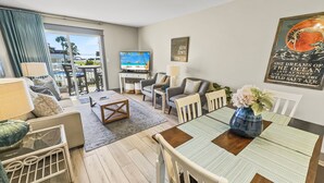 A modern living room with a large TV, comfortable seating, a dining area with a table and chairs, and a view of the outdoors. - Tradewinds 26 * Across from the beach * Fall Rates * Salt Water Vacations (Miramar Beach)