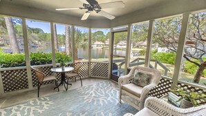 A covered patio with wicker furniture, a table, and a view of a lake and buildings.