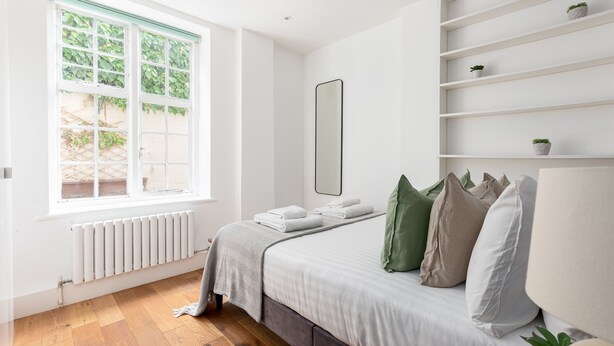 Ein Schlafzimmer mit Bett, Fenster, Heizkörper und Ablagefläche mit Pflanzen.