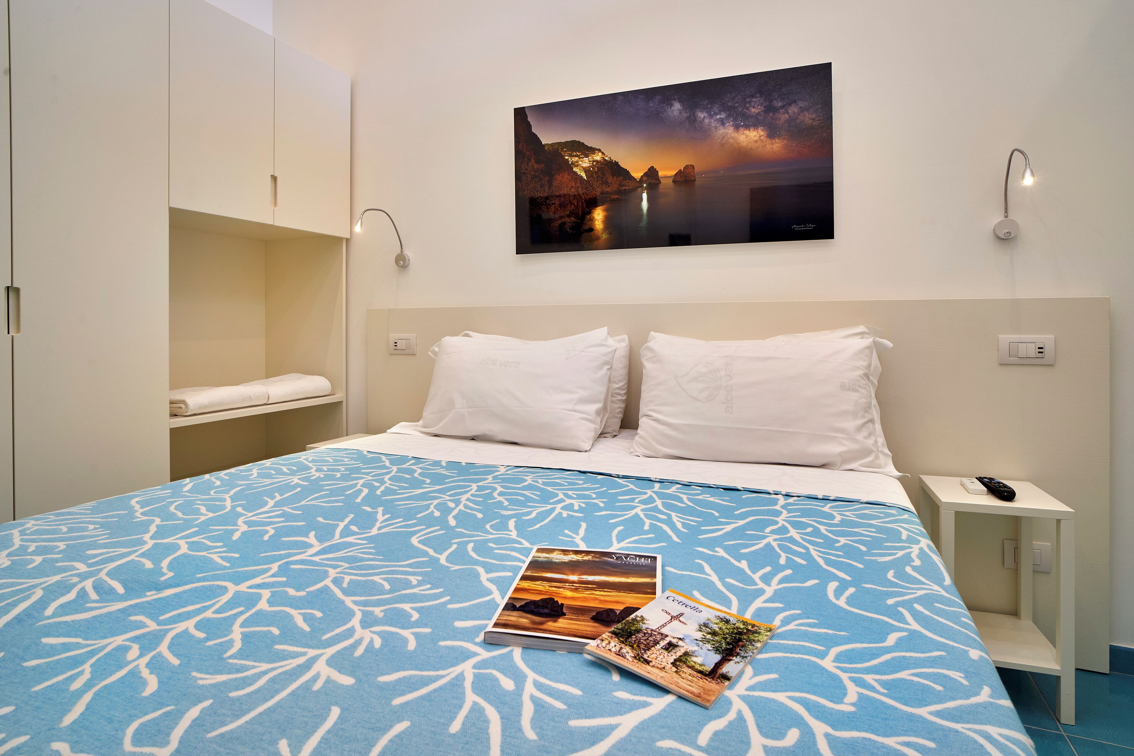 Ein Hotelzimmer mit einem großen Bett, einem Nachttisch, einem Wandbild mit Sonnenuntergang und einer Ablage mit einem gefalteten Handtuch.