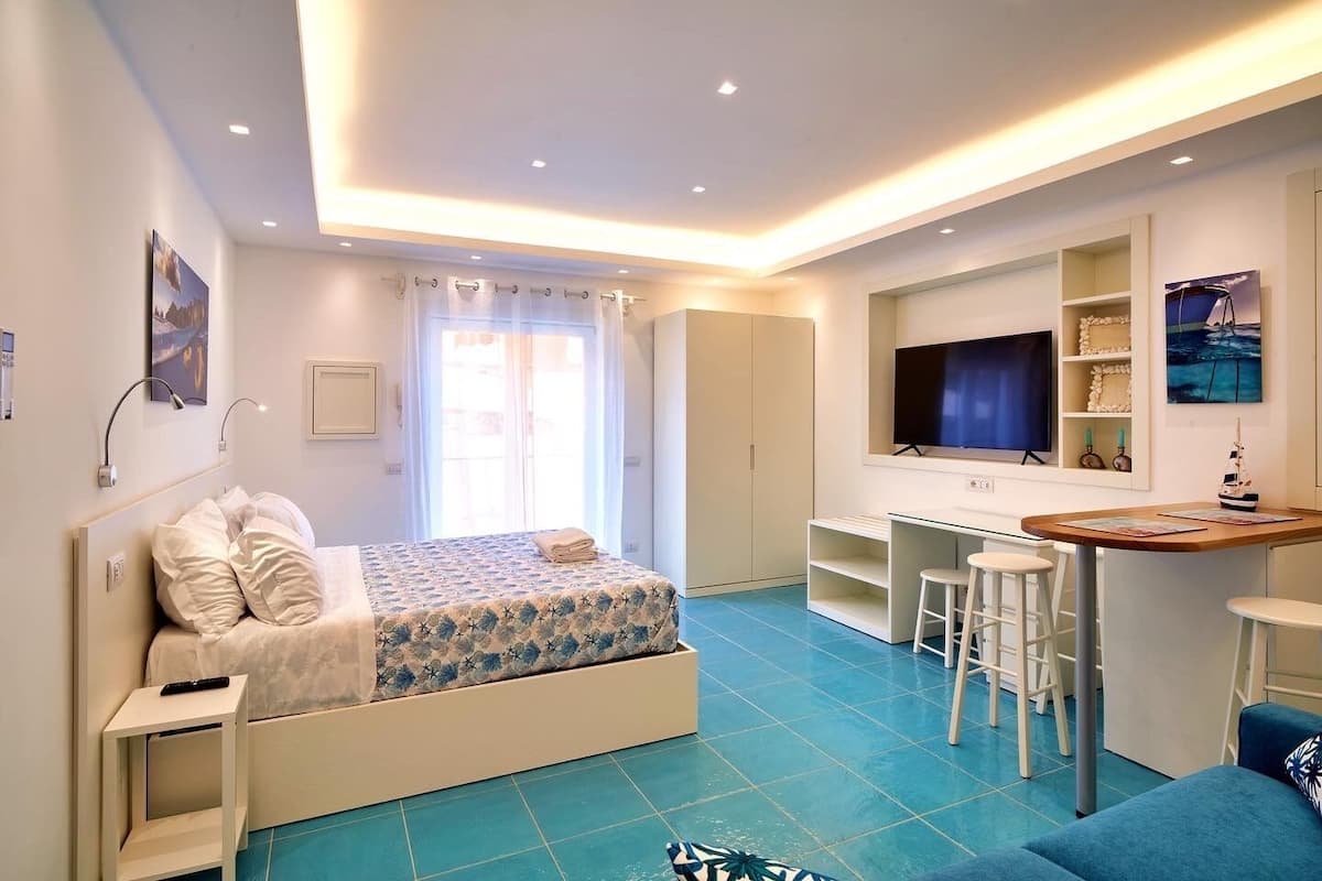 A modern bedroom with a bed, desk, and seating area, featuring a large TV and artwork on the walls.