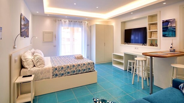 A modern bedroom with a bed, desk, and seating area, featuring a large TV and artwork on the walls. - il Truciolo Suite (Anacapri)