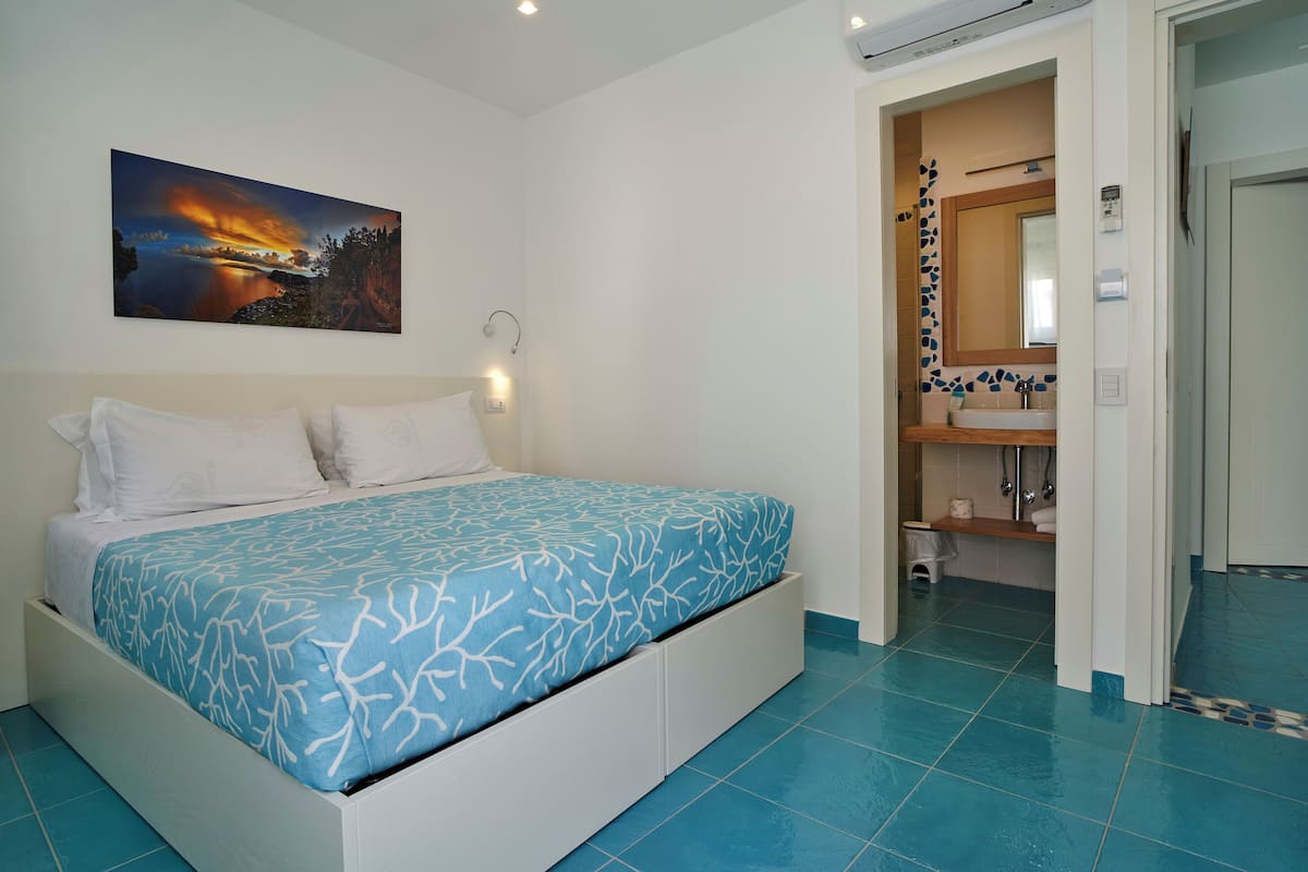 A bedroom with a bed, a nightstand, a painting, and a bathroom visible through an open door.