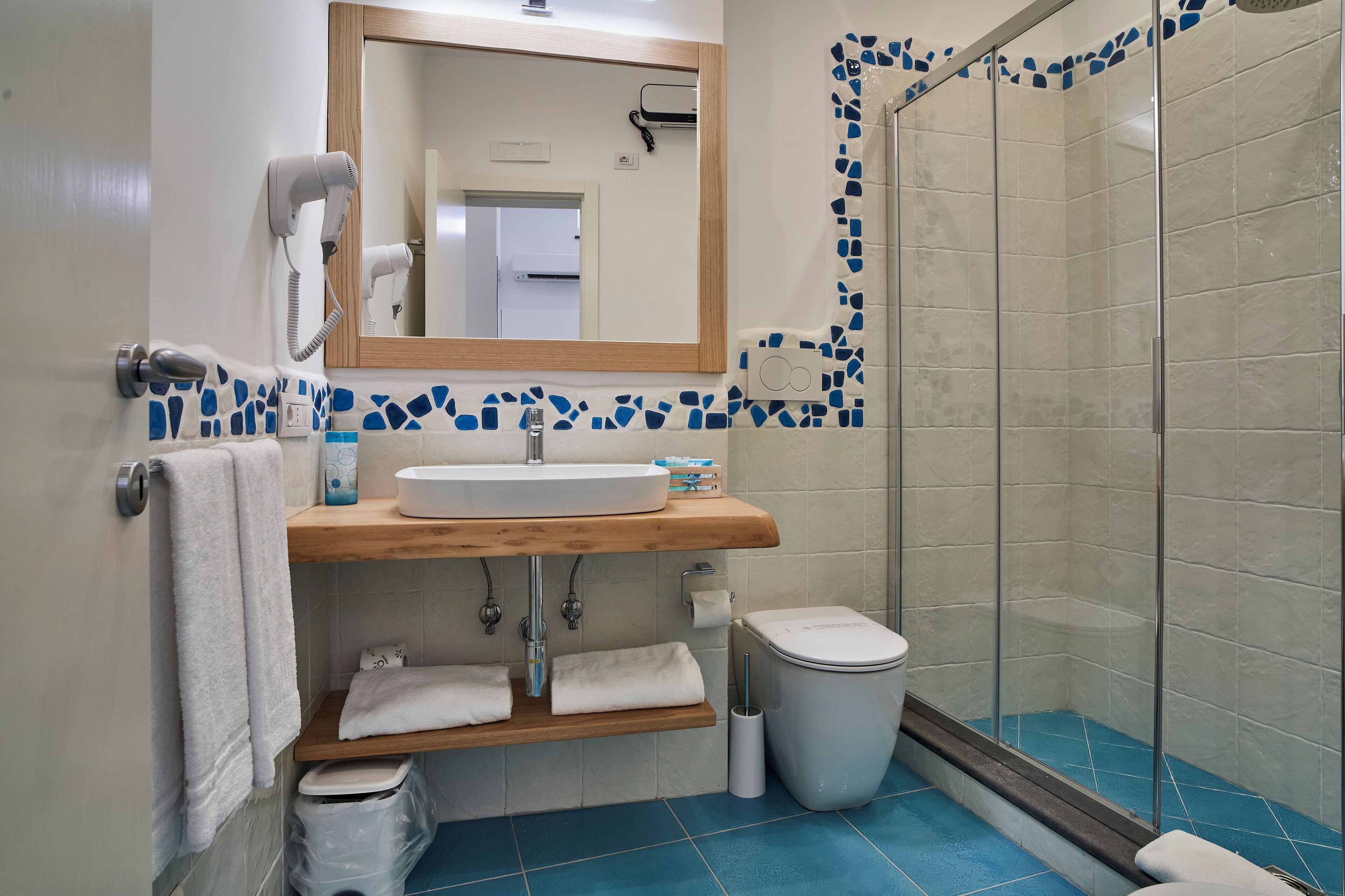 A bathroom with a wooden vanity, a mirror, a hair dryer, a toilet, and a shower area with blue tiles.