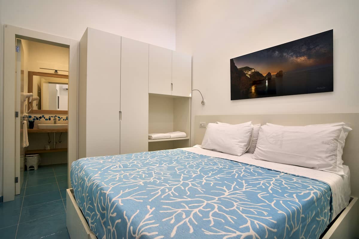 A bedroom with a bed, a nightstand, a wall-mounted painting, and a bathroom visible through an open door.