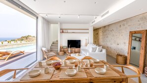 Ein modernes Wohnzimmer mit großem Fernseher, weißen Sofas und einem Essbereich mit Holztischen und -stühlen.