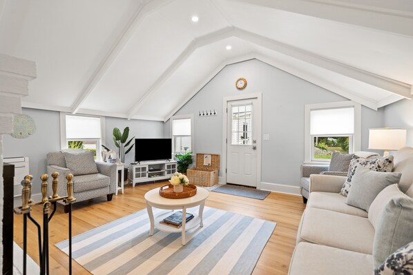 A spacious living room with a high ceiling, a flat-screen TV, a sofa, a coffee table, and a fireplace. - Shore Cottage - Coastal gem, a traveler favorite! Outdoor patio, central AC. (Gloucester)