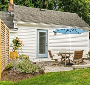 A small white house with a blue door, a patio with a table and chairs, and a blue umbrella.