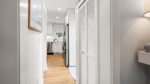 A narrow hallway with white walls and wooden flooring leading to a kitchen area with white cabinets and stainless steel appliances. - Shore Cottage - Coastal gem, a traveler favorite! Outdoor patio, central AC. (Gloucester)