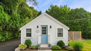 A small, single-story cottage with a blue door, a welcome sign, and a well-maintained front yard with plants and a wooden fence. - Shore Cottage - Coastal gem, a traveler favorite! Outdoor patio, central AC. (Gloucester)