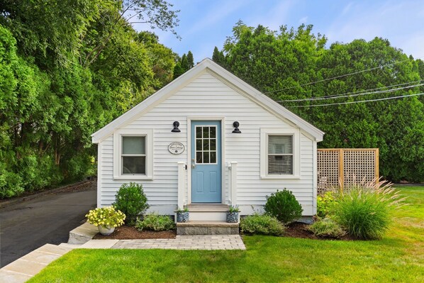 A small, single-story cottage with a blue door, a welcome sign, and a well-maintained front yard with plants and a wooden fence.