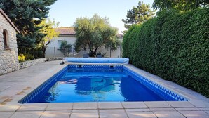 Uma piscina retangular com borda revestida em azulejo, rodeada por um pátio de pedra e um conjunto de sebe.