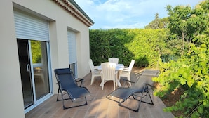 Um terraço ao ar livre com duas poltronas pretas, uma mesa e cadeiras, rodeado por um jardim e um viveiro.