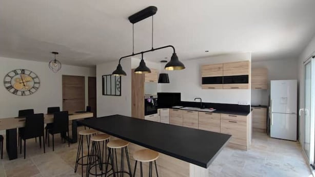 Uma cozinha moderna com uma ilha central, banquetas, iluminação pendente e um relógio na parede.