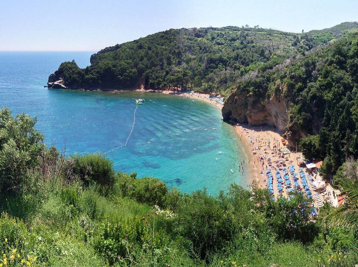 A coastal beach with clear turquoise waters, a sandy shore, and lush green hills in the background.