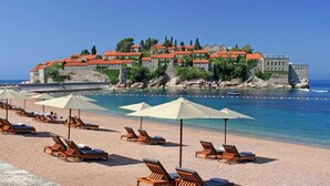 A beach with sun loungers and umbrellas, a stone building with red roofs, and a clear blue sea. - Cape Montenegro Villas (Krimovica)