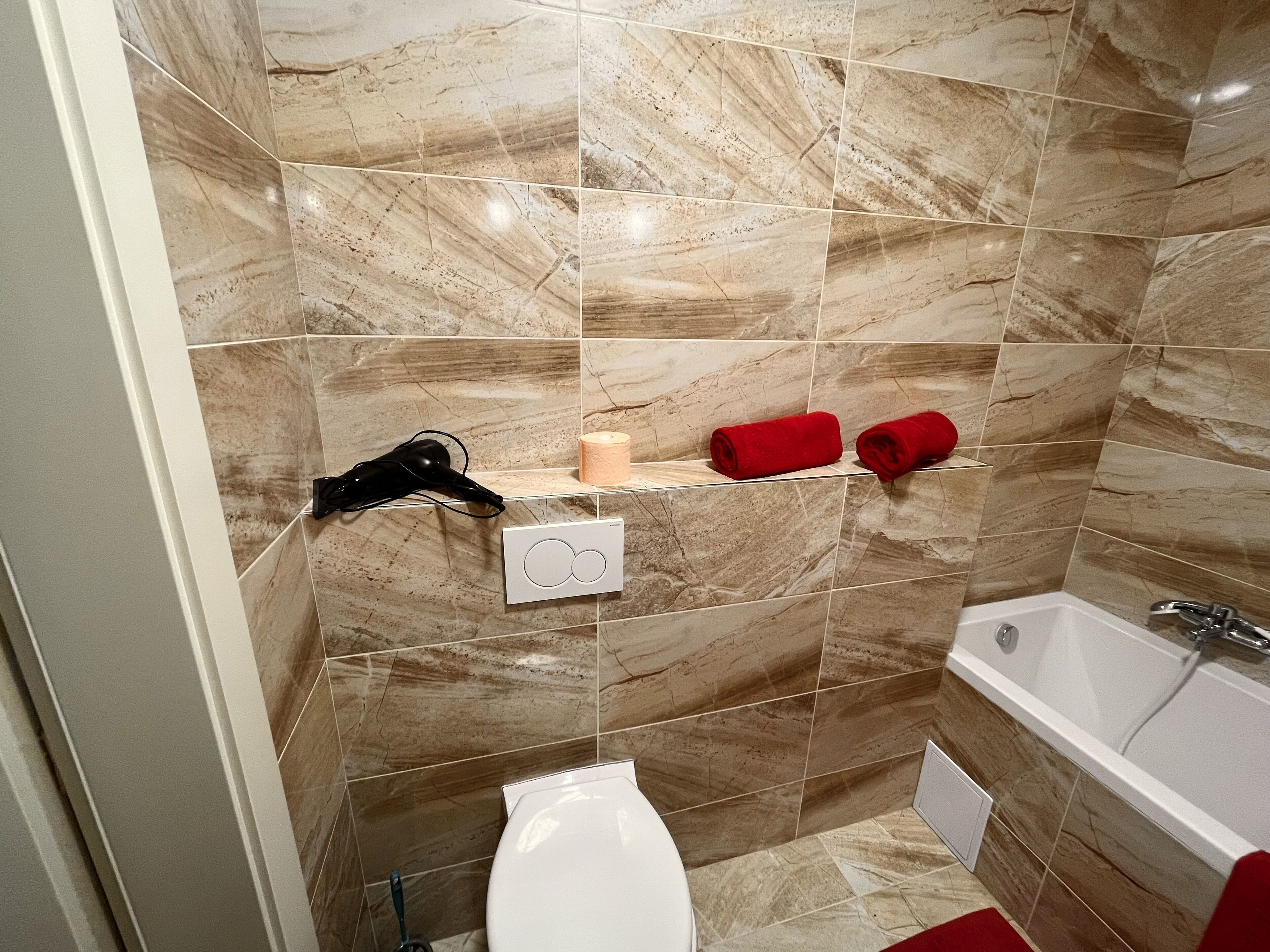 Un baño con baldosas de mármol, inodoro blanco, bañera y un perchero con dos toallas rojas.