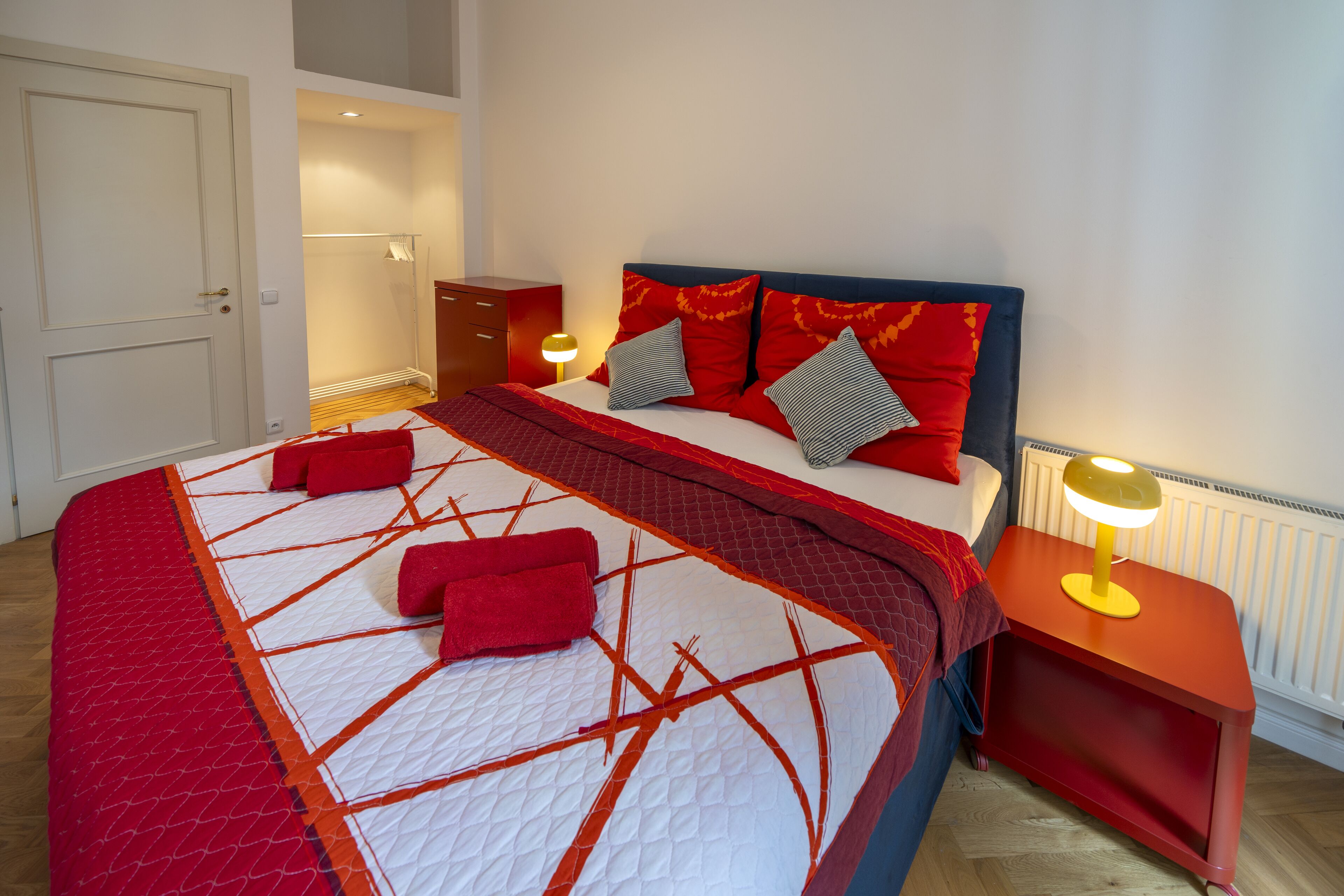 A neatly made bed with a red and white quilt, a red blanket, and several pillows. There is a bedside table with a lamp and a red nightstand.