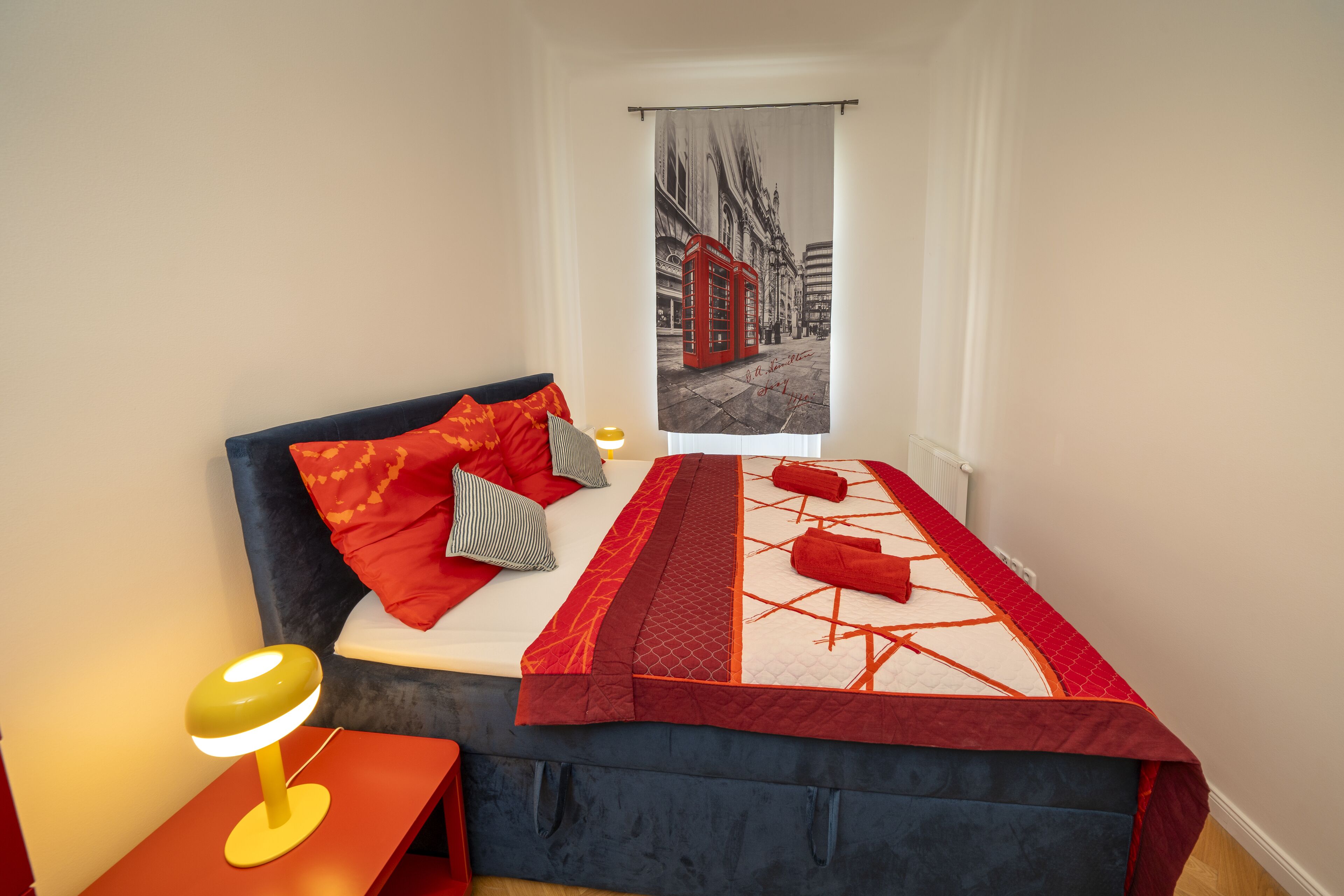 A bedroom with a bed, pillows, a bedside table with a lamp, and a view of a red telephone booth through a window.