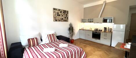 A compact bedroom with a bed, a bedside table, and a potted plant. Adjacent to the bedroom is a kitchenette with a microwave, oven, and refrigerator.