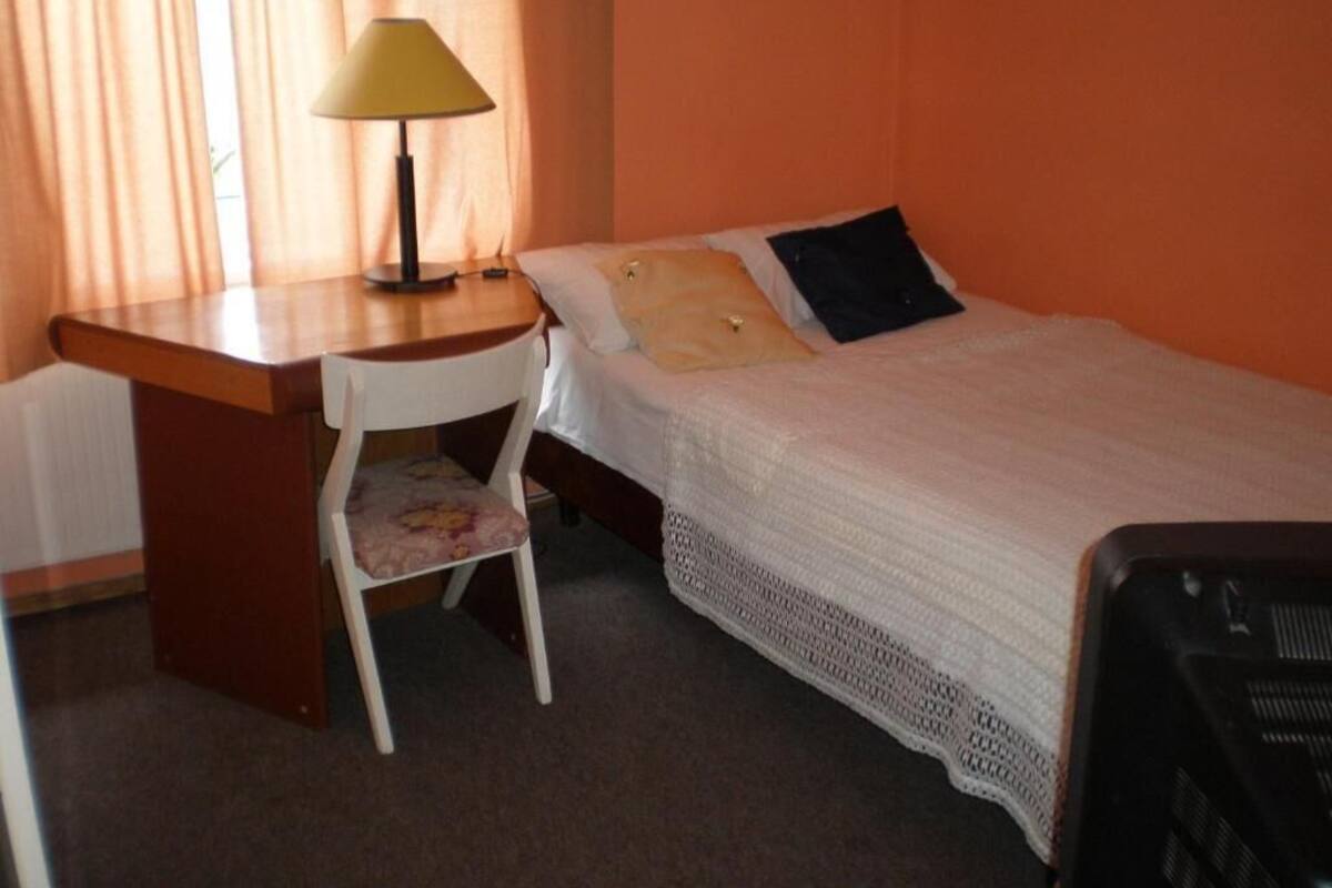 Une petite chambre d’hôtel avec un lit simple, un bureau avec une lampe, une chaise et un radiateur.