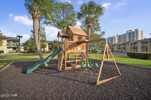 Ein Spielplatz mit Holzspielhaus, Rutschen und Schaukeln, umgeben von Palmen und einem Gebäudekomplex im Hintergrund.